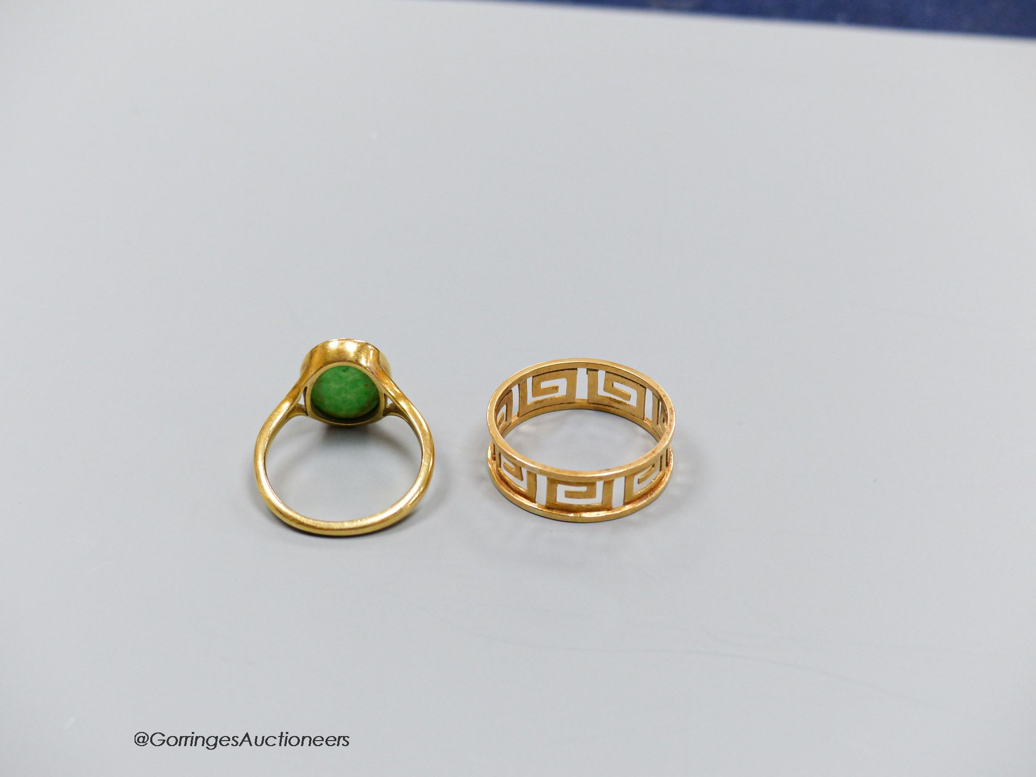 A yellow metal pierced Greek key pattern ring (testing as 18ct gold), size P, 2.2g and a similar ring set with an oval cabochon jade, size K, gross 3.2g. 5.3g